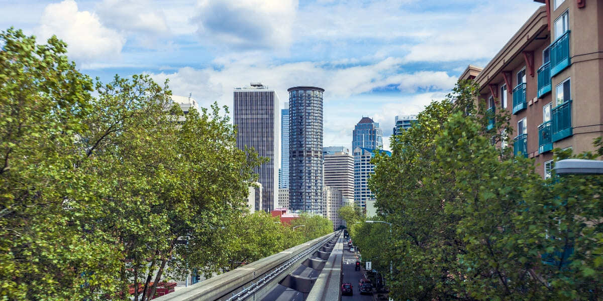 Downtown Seattle area skyline above tree-lined street and elevated light rail tracks on a bright day